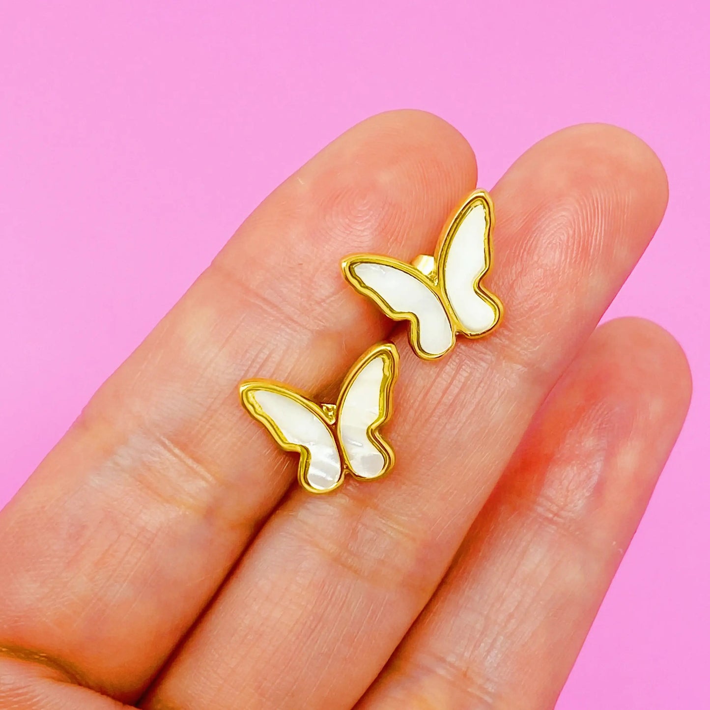 Butterfly-shaped earrings held between fingers against a pink background Close-up of two gold-edged white butterfly stud earrings delicately held between fingers with a soft pink background