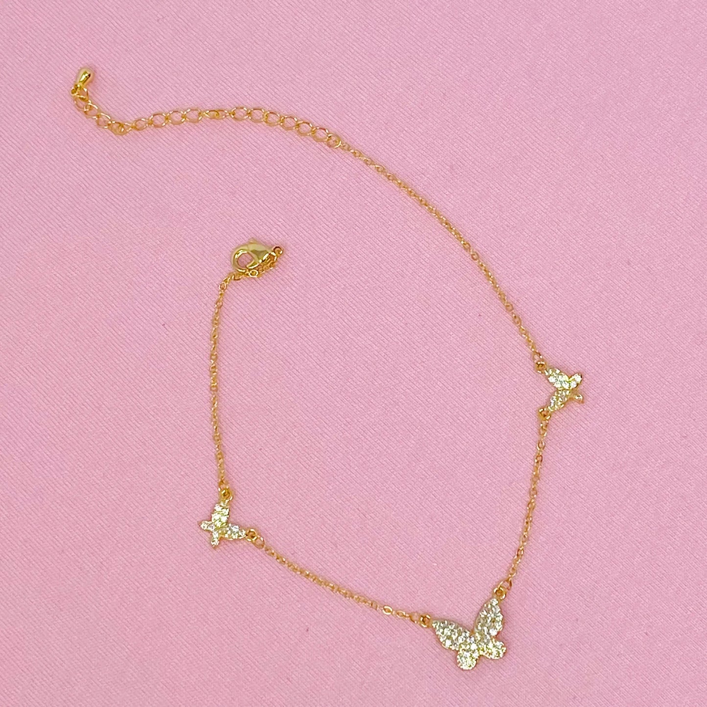 Gold necklace with green butterfly pendants on a pink background  Delicate gold necklace with three butterfly pendants displayed on a soft pink fabric background