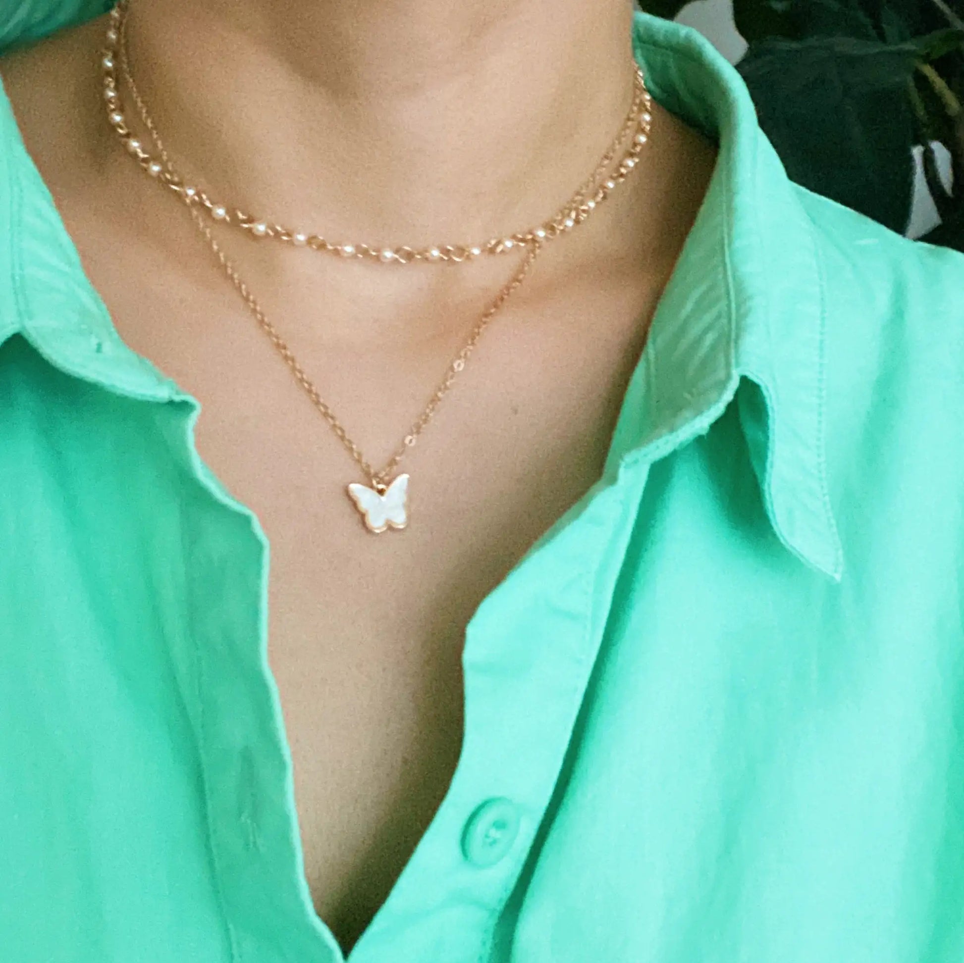 Necklace with a butterfly pendant worn by a person in a green shirt. "Layered necklace with pearl chain and gold butterfly pendant worn by a person in a green button-up shirt"
