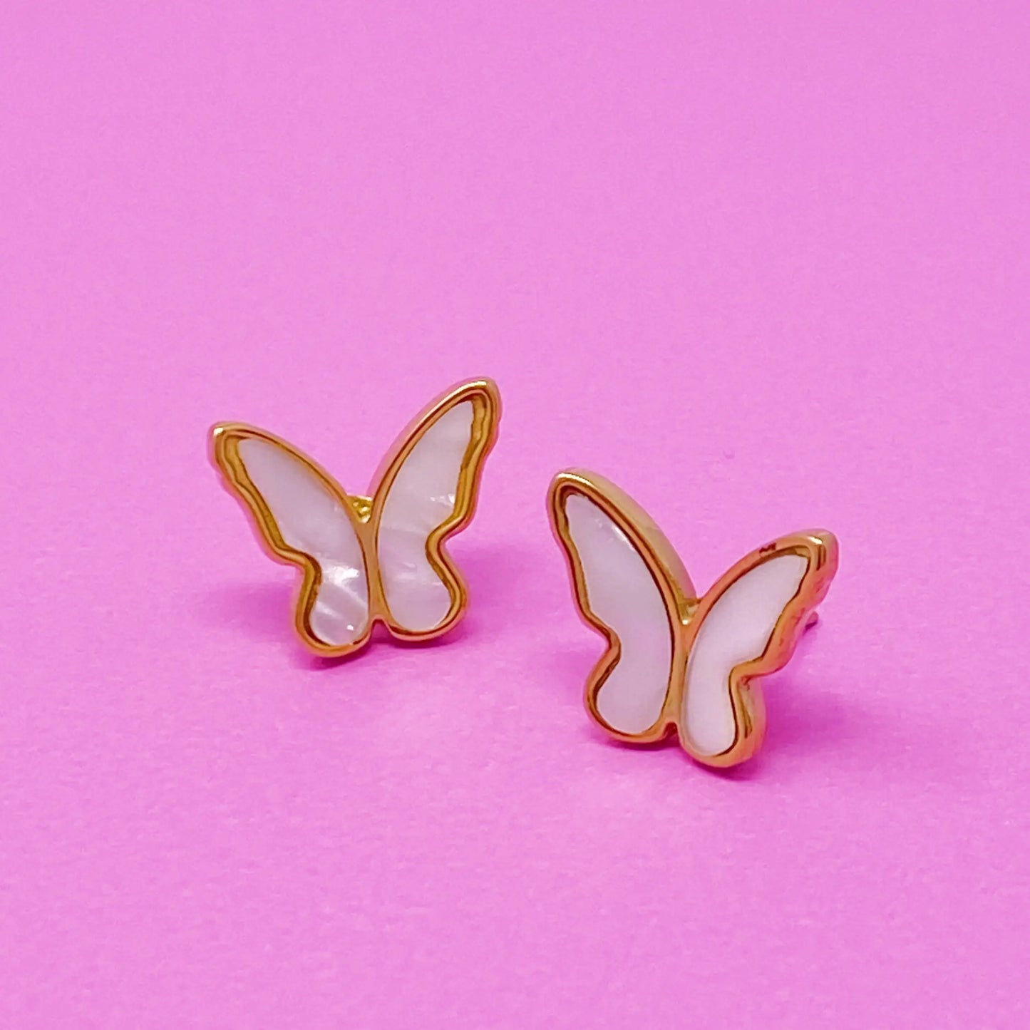 Butterfly-shaped earrings on a pink background Pair of white butterfly-shaped stud earrings with gold edges on a pastel pink background

