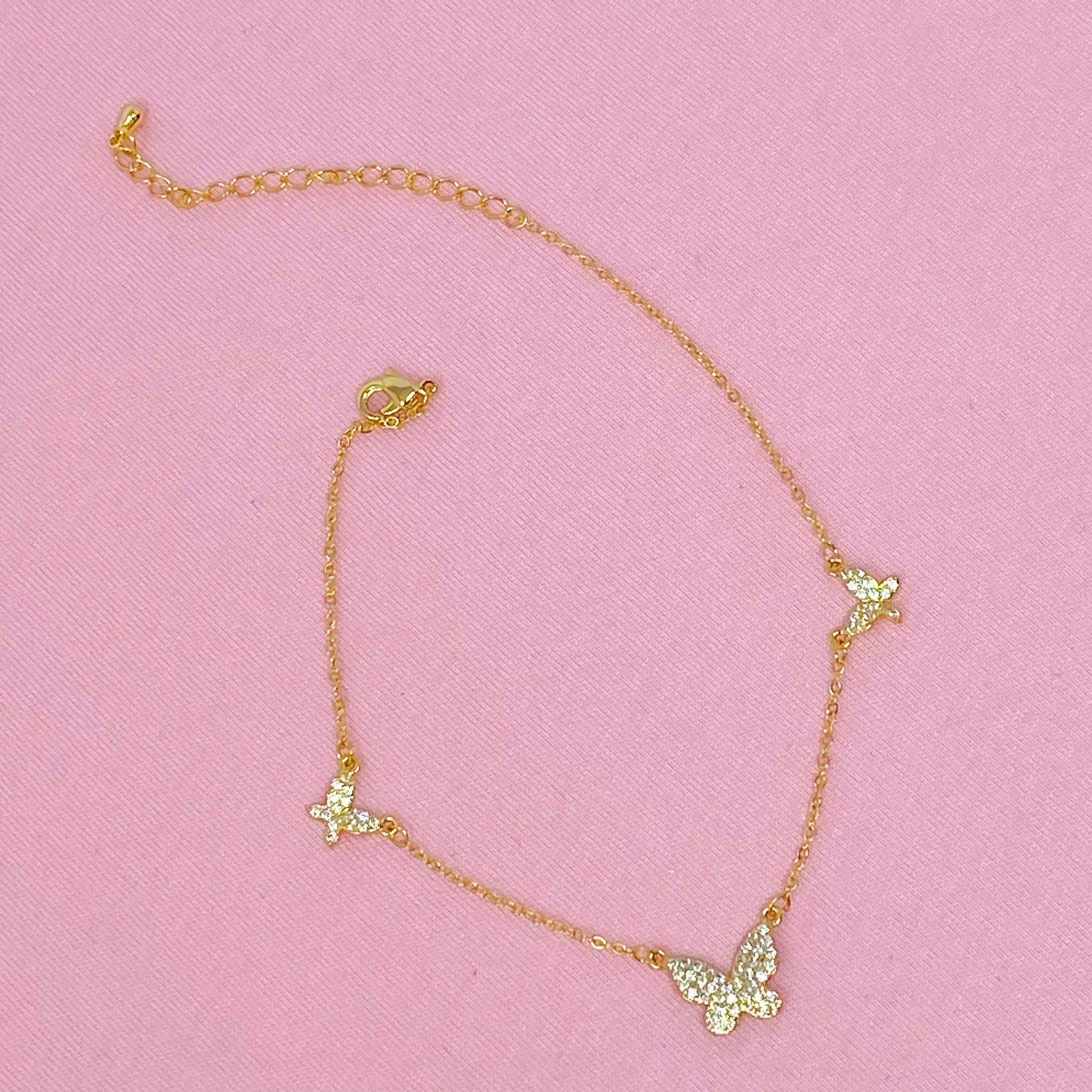 Gold necklace with green butterfly pendants on a pink background  Delicate gold necklace with three butterfly pendants displayed on a soft pink fabric background
