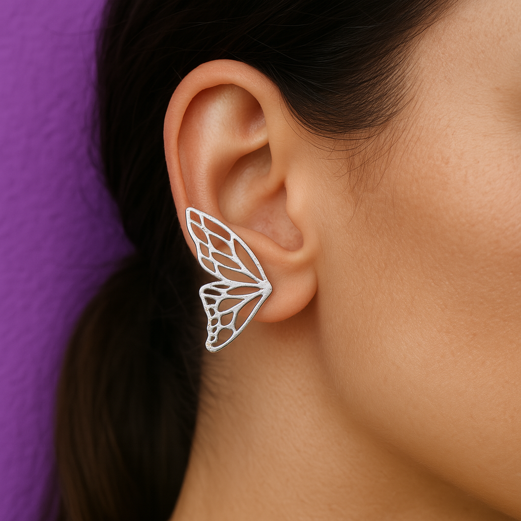 Butterfly-shaped earring worn by a person against a purple background Silver butterfly-shaped earcuff worn on a woman's ear, set against a purple background
